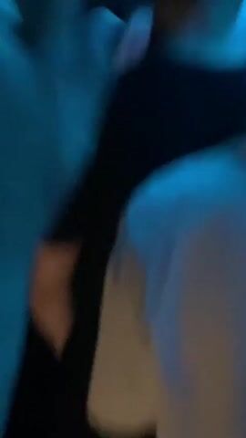 The video shows a DJ booth in a dimly lit club with blue and white lighting. A male DJ wearing a white t-shirt is in the center, operating the DJ equipment. He has tattoos on his arms and appears to be looking at the crowd. To his left, a woman in a red top is visible. The crowd is energetic, with several people raising their hands in the air and dancing. Some individuals in the foreground are blurred, suggesting they are closer to the camera. The overall atmosphere is that of a lively nightclub event.