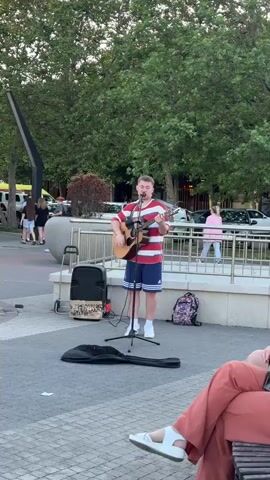 A young male street performer with short hair, wearing a red and white striped t-shirt and blue and white striped shorts, plays an acoustic guitar and sings into a microphone on a paved outdoor plaza. A black speaker with a patterned cover sits to his left, and a black guitar case lies open on the ground in front of him. A pink backpack is on the ground behind him. In the background, trees form a canopy over the plaza. Several people are walking in the distance, and parked cars are visible behind a low metal fence. A person's legs, wearing rust-colored pants and white sandals, are in the foreground, suggesting the viewer is seated on a bench or in a chair.