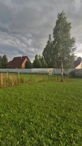 A landscape video shows a rural yard with a house and a tall tree. The sky is cloudy and appears to be darkening. The grass is green and well-maintained. There are some fences in the yard, and a greenhouse is visible in the background. The video is taken at a low angle, looking across the yard towards the house.