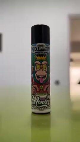 The video shows a close-up of a reusable Clipper lighter with a cartoon graphic of a "King Monkey". The monkey has swirling eyes, a crown, and holds a bowl. The background is blurry and appears to be a neutral-toned room. The lighter is placed on a light green surface.