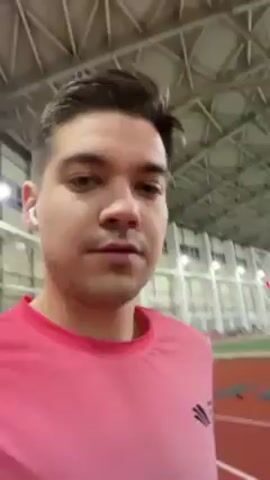 A man is walking on an indoor running track while recording a video on his mobile phone. The man has short dark hair, light skin, and is wearing a pink short-sleeved shirt with a small, dark logo on the upper left side and white wireless earbuds. He is looking directly at the camera as he walks and the camera shakes slightly as he moves. The track is red, with colored mats placed along the inner edge. The background shows an indoor sports facility with high ceilings supported by metal beams and numerous overhead lights. The facility also has large windows and some posters and banners are visible in the distance. There are other people in the background.