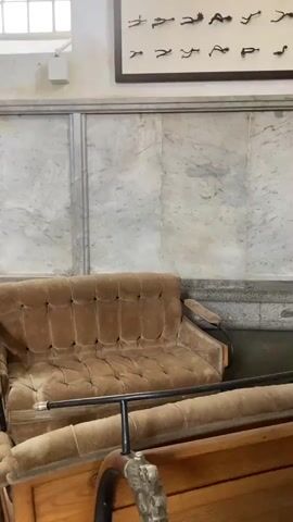 The video pans around an indoor display of carriages. The foreground shows a brown couch in a carriage and a black bar. The room has light colored marble wainscoting, framed pictures on the wall, and high windows. In the background, people stand on a balcony overlooking the carriages.