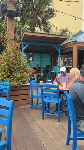 The video shows an outdoor patio area of a restaurant or bar. In the background, a man wearing a black shirt and jeans is playing an acoustic guitar and singing. He is seated in front of a teal-colored structure with a wooden roof, which appears to be a small stage or bar area. There are palm trees and string lights visible above the structure. In the foreground, several blue chairs surround wooden tables. Two people are seated at one of the tables, facing away from the camera. The person closer to the camera is wearing a white hat and a light-colored shirt. They are looking down at their hands, which are on the table. The other person has blonde hair and is wearing a grey shirt. They are also looking towards the table. The overall atmosphere appears to be casual and relaxed, with a live musical performance for diners.