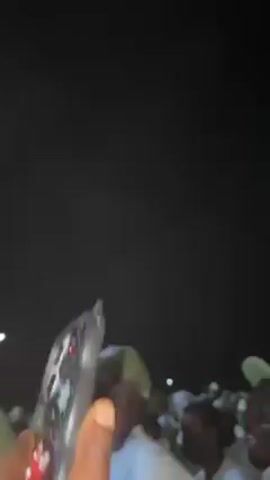 The video shows a crowd of people wearing green and white hats, likely in Nigeria. They appear to be chanting or singing loudly. The camera moves around, showing the dense crowd and a partial view of a building with brickwork. The setting appears to be outdoors at night.