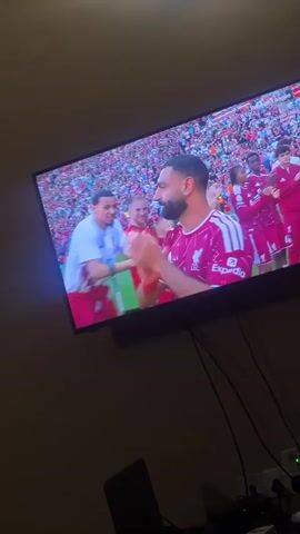 A television mounted on a wall shows a Premier League live broadcast. The screen displays footage of soccer players on a field, with a dense crowd visible in the background. A soccer player in a maroon and white jersey is being hugged. Another soccer player with long hair stands nearby, clapping. The screen then transitions to show a banner that reads "Thank You Legends" along with images of the players. The environment appears to be a residential space. The television is accompanied by several wires and a laptop on a surface below it.