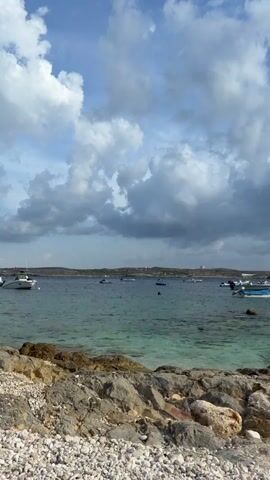 The video shows a rocky shoreline with a clear blue ocean in the background. Several boats are anchored in the water. The sky is filled with dramatic clouds, suggesting a mix of sun and possible rain. In the foreground, there are various sizes of rocks and pebbles. On the right side of the frame, a metal ladder extends from the shore into the water, indicating a possible entry point for swimming. The overall scene is a coastal landscape with a sense of tranquility, despite the dynamic sky.