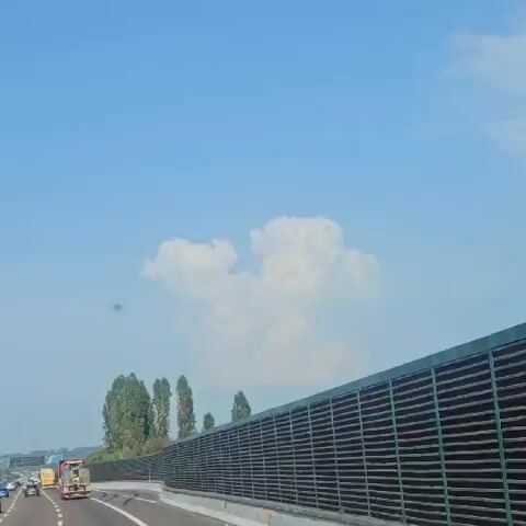 The video shows a view from a moving vehicle on a highway. A large, dark-colored sound barrier runs along the right side of the road. Beyond the sound barrier, tall trees are visible, and in the distance, a cluster of large, white clouds dominates the bright blue sky. On the highway itself, other vehicles, including a yellow truck, can be seen traveling in the same direction. The road appears to be paved with asphalt and has white lane markings.