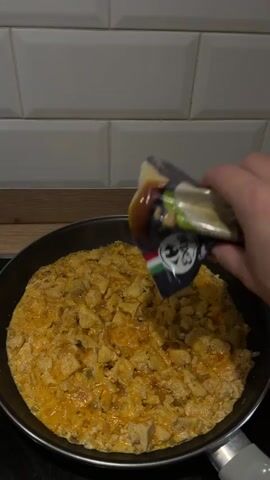 A hand is holding a small packet over a frying pan filled with a cooked dish. The packet is being tilted to sprinkle a white powder onto the food. The food appears to be chicken and eggs, with some melted cheese. The frying pan is black and sits on a stovetop. The background shows white tiled walls and a wooden countertop. The video is shot from a slightly elevated angle, looking down into the pan.