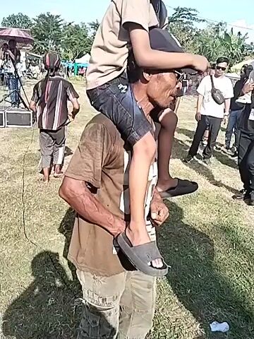 The video shows two men carrying two children on their shoulders at an outdoor event. The first man, wearing a brown t-shirt and gray slides, carries a child in a tan shirt, black jeans, and a black baseball cap. They walk on grass in front of a crowd. The second man, wearing a black shirt with a star graphic and black pants, carries a child in a white shirt and a white baseball cap. The environment is outdoors on a grassy field with people milling around, some under umbrellas, suggesting a sunny day. There are trees in the background and what appears to be a stage setup. The adults seem to be carrying the children to give them a better view of the event.