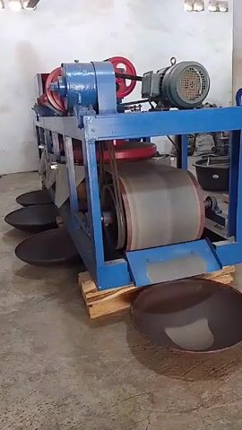 The video shows a stationary machine, primarily blue in color, with a large gray roller that spins. The machine is sitting on a wooden pallet on a concrete floor. A motor is visible on top of the machine. Several dark, round pans are placed around the base of the machine. The background shows a white wall and additional equipment.