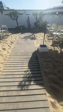 The video shows a beach with white beach huts and tables with chairs, all on a sandy surface. There is a wooden walkway leading to the huts. The sun is shining brightly, casting shadows on the sand and the walkway. A person in a dress is walking towards the huts. There are trees in white pots next to the walkway. In the background, there is a large building. Some umbrellas with blue and white stripes provide shade over some of the tables. A flag reads "STRAND". A bird flies overhead. The scene is quiet and peaceful.