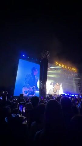 A live concert is taking place with a large crowd gathered in front of a stage. The stage is illuminated by bright lights, and a large screen displays visuals of a performer playing a guitar. Another screen shows an animated character. Above the stage, the words "PARK LIVE" are lit up in yellow. The crowd is holding up their phones to record the event. The overall atmosphere is dark, with beams of light cutting through the haze.