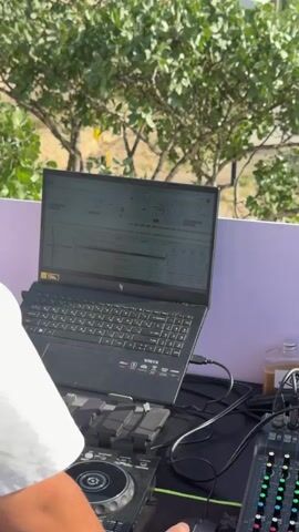 The video shows a DJ set up with a laptop and a soundboard. Behind the DJ stand, a deck with a group of people are dancing and socializing. Most are women wearing light-colored clothing. In the background, rolling green hills and orchards stretch toward a majestic mountain range with snow-capped peaks. The weather appears clear with some scattered clouds.