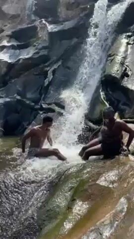 Two people are enjoying themselves in a natural pool beneath a small waterfall cascading over rocks. The person on the right is seated on a rock, while the other is in the water to the left, both enjoying the splash. The surrounding area is rocky and natural, with the focus on the waterfall and the people interacting with the water.