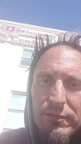 A man with disheveled hair and a goatee is standing outside of a building. He is wearing a gray hooded sweatshirt. The building behind him is a white brick structure with the words "Mission Cancer Centre" on the side. The video appears to be a selfie recording. The man is speaking.