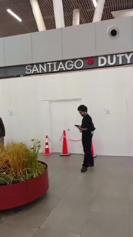 The video shows an indoor scene, likely an airport or large public building. In the foreground, there is a large red planter filled with plants, some of which are tall and grassy. To the left of the planter, a couple is standing close together, the woman embracing the man. Behind them, a sign reads "SANTIAGO". To the right of the planter, two orange traffic cones are visible, one with a rope tied between them. Further to the right, a person wearing black clothing is looking at their phone. The background shows a long white wall, closed doors, and an overhead ceiling with linear lighting fixtures. The camera pans to the left, revealing more planters with various plants, seating areas, and a woman with luggage. The scene appears to be a public waiting area, possibly for transportation.