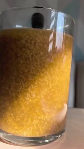 A close-up video shows a glass of a yellowish-brown liquid with a cloudy, grainy texture. The liquid is in a clear glass cup, and the cup is sitting on a light-colored surface. The background is out of focus but appears to be a room with furniture. The lighting is bright, suggesting it is daytime or a well-lit interior. The video appears to be a still shot that is slowly zooming in on the liquid. No people or other objects are clearly visible.