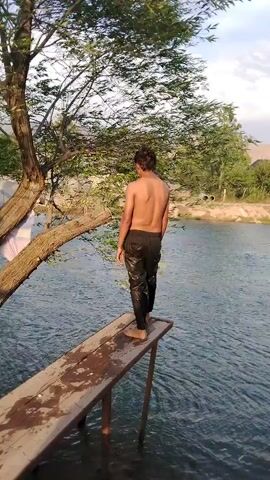 In Parchasoy, Tajikistan, a shirtless young man stands on a wooden diving board overlooking a body of water. He is wearing dark pants and appears to be preparing to jump. The diving board is elevated and extends over the water. Behind him, a large tree with green foliage is visible, its branches reaching out over the water. The water itself is calm and reflects the sky and surrounding greenery. The sky is partly cloudy, suggesting it could be daytime. The scene appears to be set in a natural, outdoor environment, possibly a river or lake. The young man's posture and focus suggest anticipation for the jump. There is no visible text in the media file.