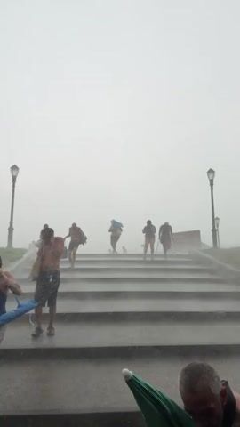 The video opens with a wide shot of a set of outdoor stairs, possibly leading up from a beach or park, on a foggy and rainy day. Several people are ascending the stairs, some carrying bags and towels. One person in the foreground, a shirtless man in shorts, is walking down the stairs. The fog obscures the background, but two lampposts are visible on either side of the top of the stairs.

The camera then pans to the right, showing a concrete wall and some vegetation.

The next scene is a close-up of a group of people gathered under an overpass, also in rainy conditions. They appear to be waiting or seeking shelter from the rain. Many are dressed in beachwear, including bikinis and swim trunks. There is a dog in a stroller, and various bags and beach accessories are visible. The overall atmosphere suggests a social gathering or the aftermath of a beach outing.