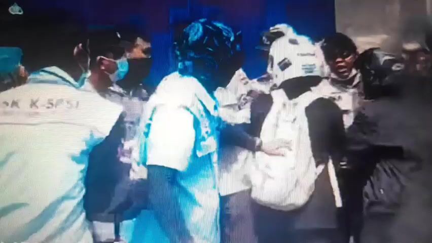 The video shows a chaotic scene with several people, some wearing uniforms and face masks, in close proximity. One individual in the foreground, wearing a white shirt, is being restrained or pushed by others. There appears to be some sort of commotion or struggle occurring. The lighting is dim, and the image quality is poor, making it difficult to discern specific details about individuals' faces or the exact nature of the interactions. The background is dark and indistinct.