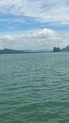 The video shows a view from a boat on the water. The water is a greenish-blue color and has small waves. In the distance, there are several islands with rocky formations. The sky is blue with some white clouds. The video pans across the scene, showing different perspectives of the islands. There are no people or specific objects visible in the video, other than the islands and the water. The overall environment is a seascape with islands.