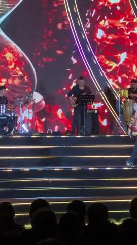 A female singer performs on a multi-level stage in Măng Đen, Vietnam. She wears a red and gold patterned jacket over dark, loose-fitting trousers. She has long dark hair and is seen walking across the stage, holding a microphone, and smiling. Behind her, a drummer plays a drum kit to the left, and a guitarist plays an electric guitar to the right.

The stage features illuminated steps with horizontal light strips. A large screen serves as a backdrop, displaying a vivid hourglass image filled with fiery red and orange patterns, creating an intense visual atmosphere. The audience is visible in silhouette at the bottom of the frame, watching the performance. The overall setting is a live concert or musical event, characterized by bright stage lighting and a dynamic visual display.