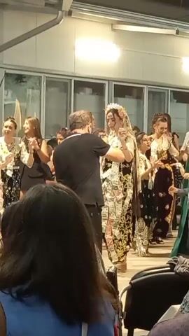 A group of people are gathered for a cultural performance. In the center, a woman adorned in traditional clothing, possibly a bride, is being presented by a man in a black shirt. Around them, other individuals, primarily women and girls, are dressed in colorful garments and appear to be participating in a dance or procession. The event is taking place indoors, likely at a community hall or similar venue, with large windows visible in the background. The audience is seated in the foreground, with their backs to the camera. The atmosphere suggests a celebratory occasion, possibly a wedding or a folk festival.