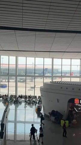 The video shows an interior view of an airport terminal. Large windows offer a view of the tarmac where several airplanes are parked. Inside the terminal, there are seating areas for passengers, and a Burger King restaurant is visible. People are seen walking around, some with luggage, and others are seated in the waiting area. The floor is highly reflective, mirroring the ceiling and the activity below. The overall atmosphere appears to be that of a busy travel hub.