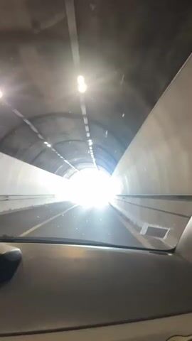 This media file captures a drive through a tunnel and out into a sunny landscape. The initial scene is inside a tunnel, with artificial lights illuminating the curved walls and ceiling. The perspective is from the driver's seat of a vehicle, looking forward through the windshield. The road ahead is visible, leading towards a bright light at the tunnel's exit.

As the vehicle emerges from the tunnel, the scene shifts to an outdoor highway. The sky is a clear, bright blue with scattered white clouds. Rolling green hills and mountains form the backdrop, with trees lining the sides of the road. The road itself is paved and appears to be a multi-lane highway with concrete barriers.

The time of day is clearly daytime, likely mid-morning to afternoon, given the bright sunlight and clear skies. The weather appears to be warm and pleasant, characteristic of a summer day.

There is a visible digital display on the lower left of the dashboard, showing "5:45" and "87". This likely indicates the time and speed of the vehicle, suggesting the drive is taking place in Italy, as indicated by the location context. The overall mood conveyed is one of movement and transition, from the confined darkness of the tunnel to the open brightness of the natural environment. No people are visible in the media file.