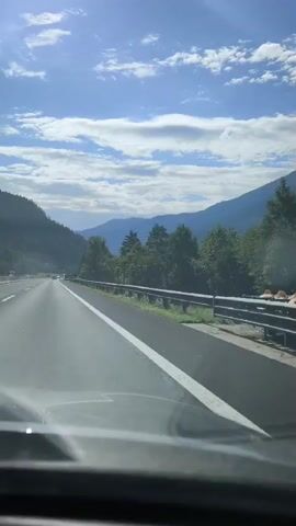 This is a first-person perspective video taken from inside a vehicle traveling on a highway. The road ahead is visible, with a white line marking the lane. On the right side of the road, there is a metal guardrail and beyond that, trees and hills. The sky is blue with scattered clouds, and the sun appears to be shining, casting some glare on the windshield. The overall environment suggests a scenic drive through a mountainous or hilly region. There are no visible people or other vehicles in the immediate foreground.