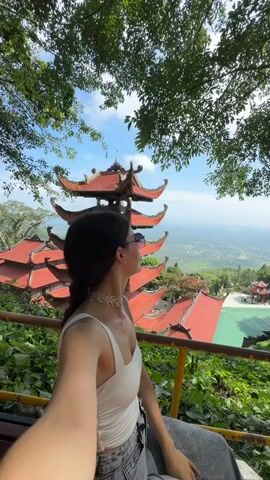 A woman with dark hair and sunglasses is sitting on a bench, taking a selfie. She is wearing a white tank top and gray jeans. Behind her is a traditional tiered temple with a red roof, surrounded by lush green foliage and trees. In the background, there is a panoramic view of a valley. The sky is blue with some clouds. The camera pans across the temple and the surrounding landscape.