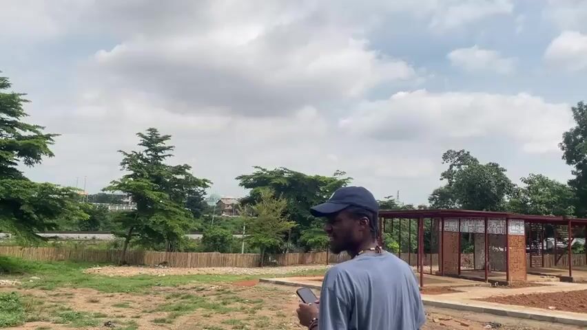 The video captures a moment in an outdoor setting, possibly in Wupa, Nigeria. The primary subject is a young Black man wearing a blue baseball cap and a grey t-shirt. He is seen in the foreground, looking away from the camera and holding a dark object, likely a phone, in his right hand. He appears to be engaged in some form of contemplation or action with the object.

The scene is an open, somewhat undeveloped area. The ground is a mix of dirt and patchy grass. In the mid-ground, a low wooden fence made of natural bamboo-like material encloses a section of the area. Beyond the fence, lush green trees of varying sizes are prominent, creating a natural backdrop. One particularly tall, conical evergreen tree stands out to the left. In the distance, more trees and some structures can be seen, hinting at a developing area or a community. To the right of the frame, there is a partially constructed open-air structure with a red roof, supported by metal beams and featuring brickwork.

The sky is filled with dramatic, billowing white clouds against a pale blue background, suggesting it is daytime, likely late morning or afternoon. The lighting is somewhat diffused, indicating overcast conditions or a less direct sunlight. The overall atmosphere is relaxed and casual. There is no visible text in the video. The setting appears to be a public space undergoing development, with a blend of natural elements and construction.