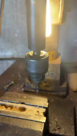 The video shows a close-up of a drill press in operation. The drill is spinning and being lowered into a metal workpiece held in a vise. Metal shavings are visible around the work area. The machine is well-lit, likely with a built-in lamp. The overall setting appears to be a workshop or garage.