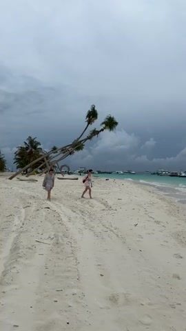 Two women are walking toward the camera on a white sandy beach on a cloudy day. They are both wearing light-colored summer clothes. One has a red bag on her shoulder. Behind them are several palm trees, some of which are leaning. In the distance, there is turquoise water with several boats docked. There are marks on the sand from vehicle tires.