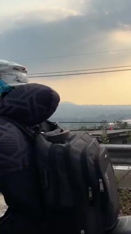 The video shows a view from a road overlooking a valley. In the foreground, there is a gray concrete barrier with a green metal railing. Beyond the railing, there is a construction site with rebar and concrete structures. Further in the distance, there are trees, fields, and a body of water. The sky is cloudy with hints of sunset colors. The camera pans slowly to the right, revealing more of the landscape and a papaya tree with a white painted trunk. The overall impression is one of a developing area with natural scenery in the background.