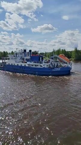 A blue and white tugboat, named "VESELAYA SAMRA" is shown moving along a wide, brown-colored river under a sunny sky with scattered clouds. The tugboat is pulling a long, dark barge. The video is shot from the side of another boat, with the white railing of that boat visible in the lower left corner of the frame. The river is bordered by green trees on either side. The tugboat has a boxy shape with a deckhouse, an orange life raft, and various antennas and structures on its top.