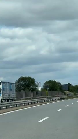 The video shows a view from a moving vehicle on a highway. There are several cars and trucks on the road. On the side of the highway, there is a concrete barrier, trees, and vegetation. In the background, there are some industrial buildings with tall structures, and a large transmission tower. The sky is cloudy.