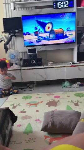 A young child, a boy wearing a striped t-shirt and patterned shorts, sits on a light-colored mat with cartoon drawings. He is looking down and appears to be eating something from his hand. The child is in a living room with a large flat-screen television displaying a cartoon. The television is mounted above a white media console with speakers and other electronic devices. A blue fan is visible to the left of the television. The room has tiled flooring and a white wall with some shelving. The lighting suggests it is daytime.