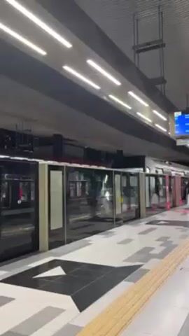 A white train with black window frames is seen moving through a station. The train is traveling from left to right in the frame. The platform has patterned black and gray tiles. Rows of overhead lights are visible in the ceiling. A yellow tactile strip is visible along the edge of the platform. Some people can be seen inside the train as it travels by.
