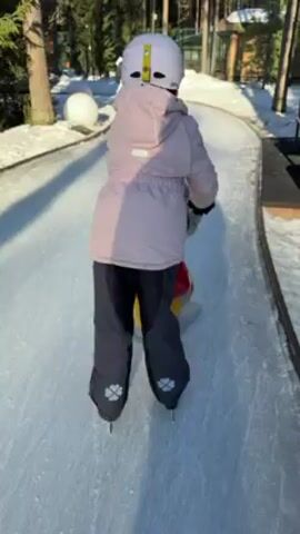A young child, viewed from behind, is ice skating on a prepared ice path. They wear a white helmet with a yellow stripe, a light pink jacket, and dark blue pants adorned with gray four-leaf clover designs on each ankle. They are assisted by a colorful ice-skating aid. The ice path is well-lit and surrounded by trees and snow. To the side of the path is a dark brown wooden structure. Additional elements in the background include white spherical decorations and a building in the distance. The child appears to be moving forward along the ice path.