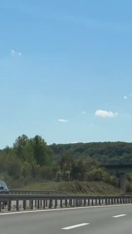 A car is driving down a highway on a clear day. The highway is surrounded by trees and hills. A bridge is visible over the highway. There are other cars on the highway.