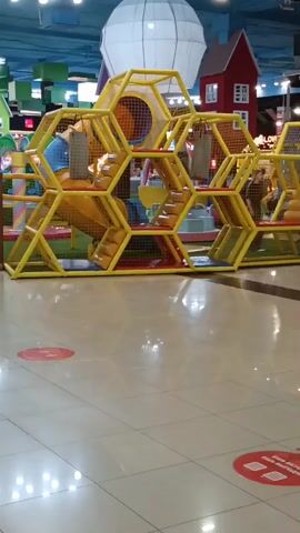 The video shows an indoor playground with a honeycomb-shaped structure. Yellow hexagonal frames form the structure, with orange padded sections and yellow slides. Several children are visible playing on the structure, climbing, sliding, and interacting. The playground appears to be in a shopping mall or similar public space, with tiled flooring and a large white balloon-like object in the background, along with small decorative houses. Red circular markers are visible on the floor, indicating social distancing guidelines. The lighting is bright, suggesting an indoor artificial lighting setup.