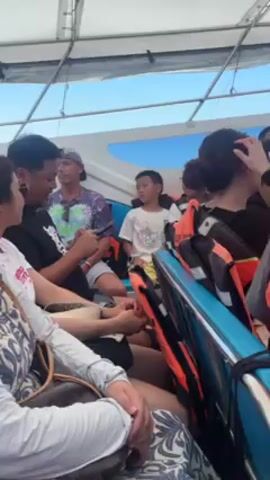 The video shows the interior of a moving boat on a sunny day. Several people are seated on the blue seats. Some are wearing life jackets.  A man in a black shirt is looking at his phone. A young boy in a white t-shirt sits next to another boy with glasses.  A woman in a white patterned top sits in the foreground.  The roof of the boat is visible above, and the sky is seen through a window at the front.