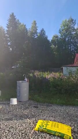 The video shows an outdoor scene with a man and several objects. In the foreground, there is a bag of green material on a gravel surface. A white plastic table with a plastic tablecloth is to the left, with some items on it. A large gray metal barrel with a faucet and a white bucket at its base is in the middle of the frame. Further back, a blue inflatable pool is visible on the grass to the left of the table. A green inflatable object, possibly a boat or mattress, is lying on the grass to the right. In the middle ground, a shirtless man wearing dark shorts is standing near a grill that is emitting smoke. He appears to be tending to the grill. Behind the grill, there are wooden structures, one of which appears to be a building or shed. Trees and vegetation, including a patch of purple flowers, are visible in the background. The sun is shining brightly, creating lens flare and casting shadows. The sky is blue with some clouds.