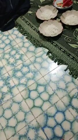 A video shows two individuals preparing food on the floor. The floor is tiled with a hexagonal pattern in blue and green. A patterned rug with a fringed edge is spread on the floor. Several plates are arranged on the rug, each containing white rice. Some plates also have slices of watermelon. One person, wearing a dark top and a blue headscarf, is sitting on the floor and appears to be serving food from a bowl. Another person, wearing a striped shirt and a dark blue garment, is also sitting on the floor and is partially visible in the frame. In the background, there are cabinets, a pink bowl, and some red items on a plate, possibly more watermelon or other food. The overall scene suggests a gathering or a meal preparation.