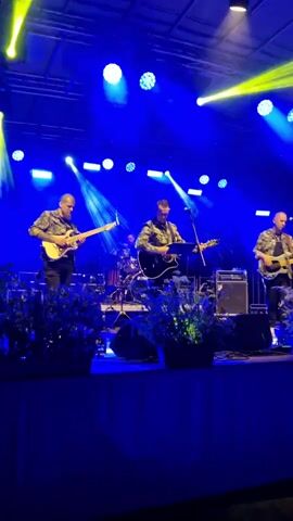 A band performs on a stage illuminated by blue and yellow lights. There are three guitarists in the foreground, all wearing camouflage shirts. The guitarist on the left is playing a white electric guitar. The guitarist in the center is playing an acoustic guitar and singing into a microphone. The guitarist on the right is playing a white electric guitar. In the background, a drummer is visible behind a drum set. There are potted plants on the edge of the stage in front of the band.