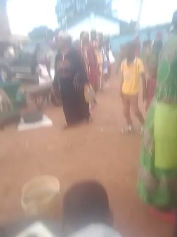 The video shows a group of people, predominantly children, standing in a line outdoors on a dirt surface. Some adults are also present, seemingly supervising the children. The children are dressed in various colorful clothing, including red, yellow, and blue outfits. Some of the children are wearing shoes, while others appear to be barefoot. The environment suggests a casual gathering or event, possibly a school activity or a community function. There are some objects visible in the background, including a building with a light-colored wall and a dark roof, and some trees. A plastic bucket is also visible near the front of the group. The lighting indicates it is daytime, with the sun casting shadows on the ground. The camera is held at an angle, giving a slightly distorted perspective of the scene.
