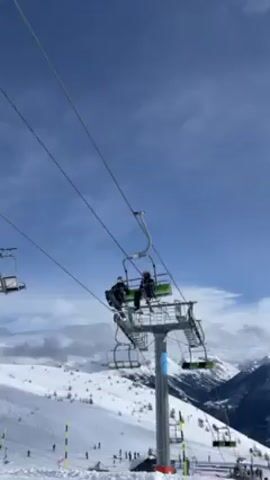 The video shows a ski lift moving upwards through a snowy mountain landscape under a partly cloudy sky. Two people are riding on one of the chairs, one with skis and the other with a snowboard. The ski lift structure is visible, including the cables, towers, and empty chairs. In the background, there is a snow-covered mountainside with skiers and snowboarders visible on the slopes. The person filming is wearing a ski helmet.