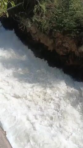 A high-angle shot looks down at a powerful waterfall cascading over rocks. The water is white and frothy, creating a turbulent surface. Lush green trees and brown rock formations line the top of the waterfall. The bottom of the frame shows a concrete edge.