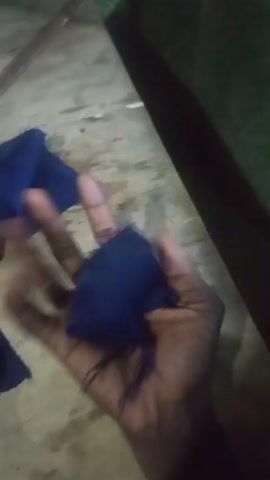 The media file shows a close-up of a person's hands manipulating dark blue fabric. The hands are dark-skinned. The fabric appears to be a rough, woven material, possibly cotton or a blend. In the background, a wooden surface with stains and debris is visible, suggesting a workspace or a casual setting. To the right of the frame, a dark green object, possibly furniture or a large piece of equipment, is partially visible. There are also other pieces of fabric scattered around, including one with stripes of pink, green, and white. The lighting is dim, with some areas brightly illuminated and others in shadow, indicating an indoor environment with artificial light, or it could be at night. No text is visible in the media file. The activity appears to be related to sewing, crafting, or preparing fabric for some purpose. There are no clear indicators of the time of day or weather. The focus is on the tactile interaction with the fabric.